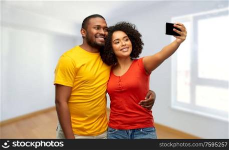new home, real estate and people concept - happy smiling african american couple taking selfie by smartphone over empty apartment on background. african american couple takes selfie by smartphone