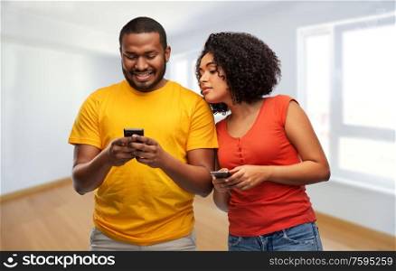 new home, technology and people concept - happy african american couple with smartphones over empty apartment on background. happy african american couple with smartphones