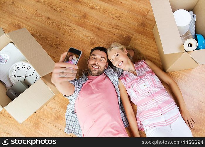 new home, technology, people, repair and moving concept - happy couple taking selfie with smartphone and lying on floor among cardboard boxes at home