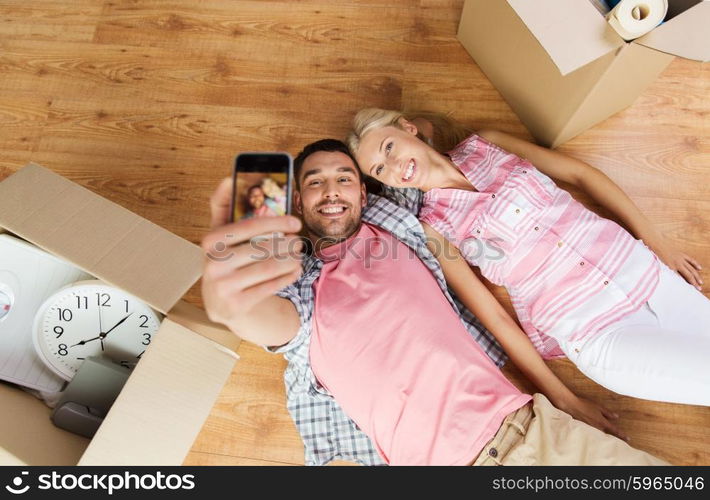 new home, technology, people, repair and moving concept - happy couple taking selfie with smartphone and lying on floor among cardboard boxes at home