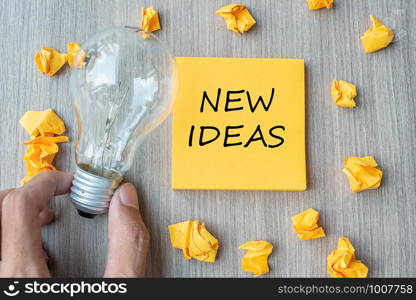 New Idea words on yellow note and crumbled paper with Businessman holding lightbulb on wooden table background. Creative, Innovation, Imagination, inspiration, Solution, Strategy and Goal concept