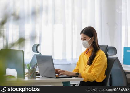 New normal of Asian woman in yellow shirt wearing surgical face mask working with computer laptop thinking to get ideas and requirement in Business startup at modern office or Co-working space