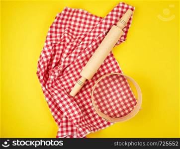 new wooden rolling pin on a red textile napkin and a round sieve for flour, yellow background, top view