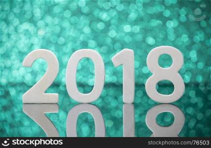 New year 2018 wood numbers reflexion on glass table in front of white lights bokeh over green background.