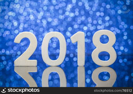 New year 2018 wood numbers reflexion on glass table in front of white lights bokeh over blue background.