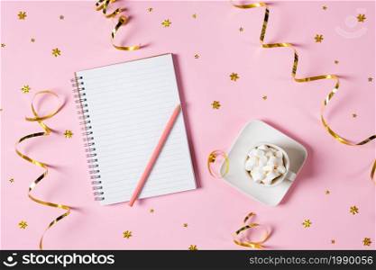 New year, Christmas or holiday wish list concept. Notepad, golden tinsel, and a cup of hot chocolate with marshmallows isolated on pink background. Holiday banner with copy space.. New year, Christmas or holiday wish list concept. Notepad, golden tinsel, and a cup of hot chocolate with marshmallows isolated on pink background. Holiday banner with copy space