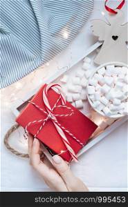 New Year or Christmas flat lay top view with Hot cacao coffee chocolate with marshmallows mug Xmas holiday celebration red present box on wooden tray in bed with lights and woman hands. Concept 2020