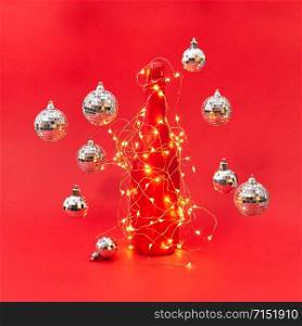 New Year shined lights string on a creative painted wine bottle and floating balls around on a red background with copy space. Christmas congratulation card.. Wine bottle painted red covered garland with lights and flying balls.