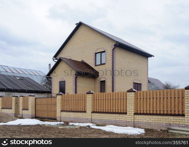 New yellow brick cottage behind a high fence in early spring