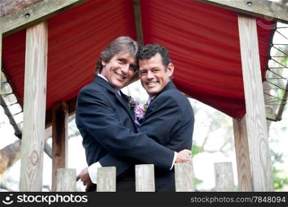 Newly married gay couple embracing under an outdoor canopy.