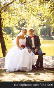 Newly married smiling couple sitting on bench under tree at river bank