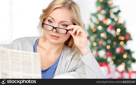 news, press, media, holidays and people concept - woman in eyeglasses reading newspaper at home over christmas tree lights background