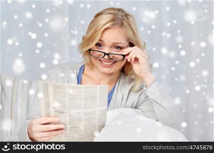 news, press, media, leisure and people concept - woman in eyeglasses reading newspaper at home over snow