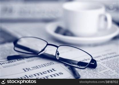 Newspapers and coffee cup, with reading glasses. Toned image, focus on reading glasses.