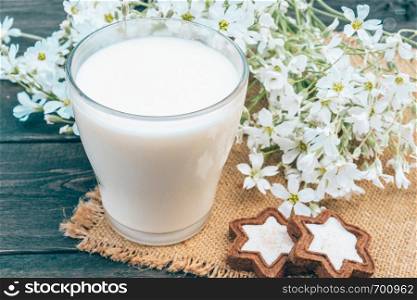 Next to the cookies and small white flowers is a glass cup of milk on a napkin.. Next to the cookie and white flowers is a glass cup of milk on a napkin.