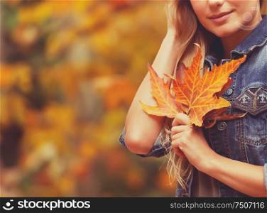 Nice female over autumnal foliage background with beautiful dry maple leaf in hand, autumn vacation concept