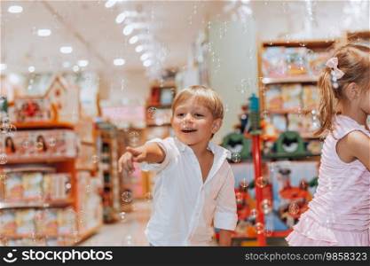 Nice little brother and sister with pleasure blowing soap bubbles in a toy store. Cheerful kids having fun in the child’s room. Happiness all around.. Happy Children Blowing Soap Bubbles
