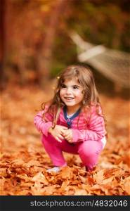 Nice little girl playing in autumn park, sweet kid sitting on the dry brown tree leaves, enjoying beauty of the fall