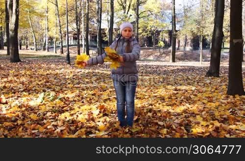 nice teen girl rotates and scatters yellow leaves in beautiful autumn city park
