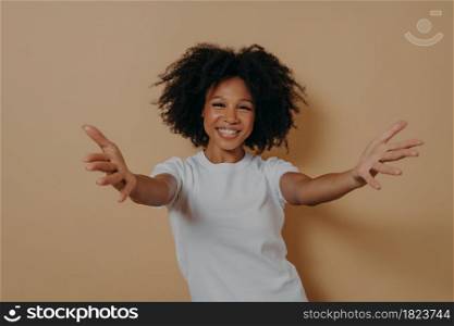 Nice to meet you. Overjoyed happy african woman 20s standing isolated on beige background with hands wide open, kind positive female in white tshirt wants to hug person. Invitation concept. Overjoyed happy african woman wants to hug person while standing isolated on beige background