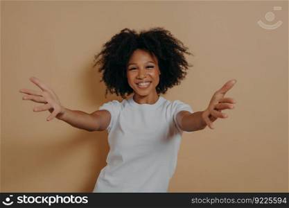 Nice to meet you. Overjoyed happy african woman 20s standing isolated on beige background with hands wide open, kind positive female in white tshirt wants to hug person. Invitation concept. Overjoyed happy african woman wants to hug person while standing isolated on beige background
