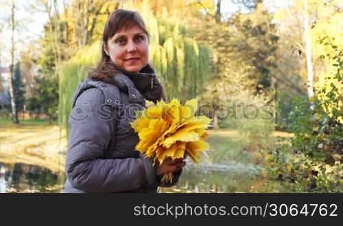 nice woman sits and holds in her hands yellow leaves at beautiful autumn city park near lake, closeup