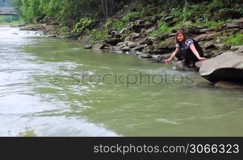 nice woman sits and plays with water on shore of mountain river