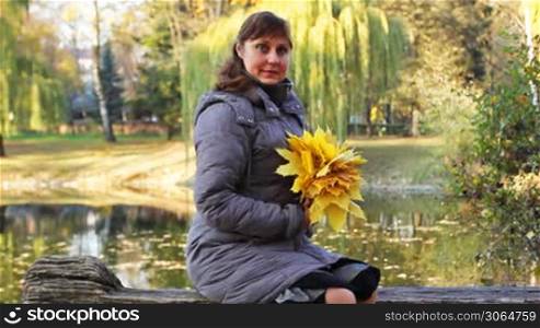 nice woman sits on bench and holds in her hands yellow leaves at beautiful autumn city park near lake