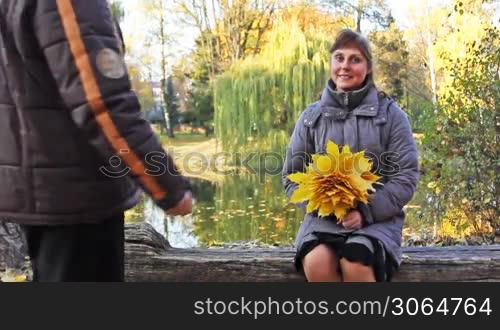 nice woman sits on bench, to her approaches man in beautiful autumn city park near lake
