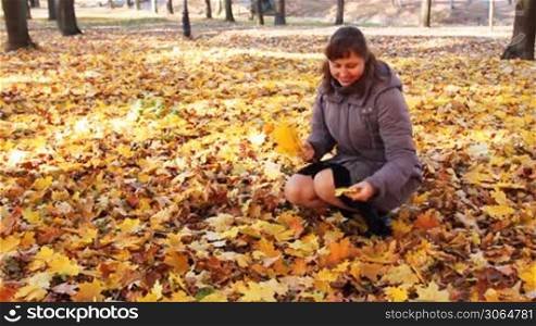 nice woman sitting squatted collects yellow leaves and smiles in beautiful autumn city park