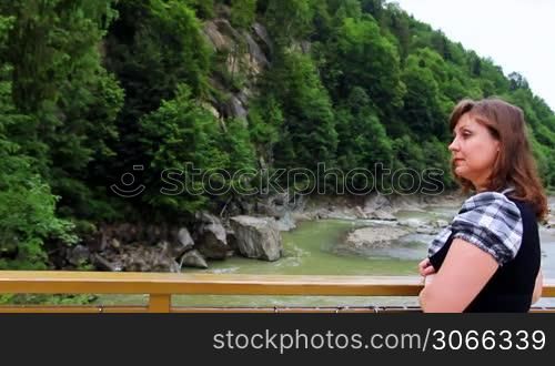 nice woman stands near railing bridge, then looks on waterfall in Carpathian Mountains