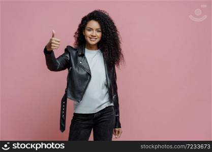 Nice work! Pleased curly haired woman keeps thumb up, makes approval gesture, dressed in white t shirt, stylish leather jacket and jeans, poses against pink background. Body language concept