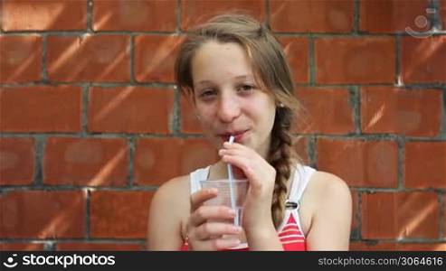 nice young girl sits in front of brick walls and drinks from plastic water cup through straw