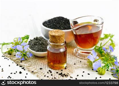 Nigella sativa oil in vial and gravy boat, seeds in a spoon and black cumin flour in a bowl on burlap, kalingi twigs with blue flowers and leaves on light wooden board background