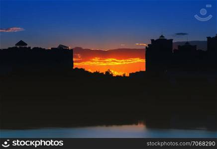 Night city landscape - silhouettes of high-rise buildings against the background of an orange sunset reflected in the lake.. Night City Landscape At Sunset