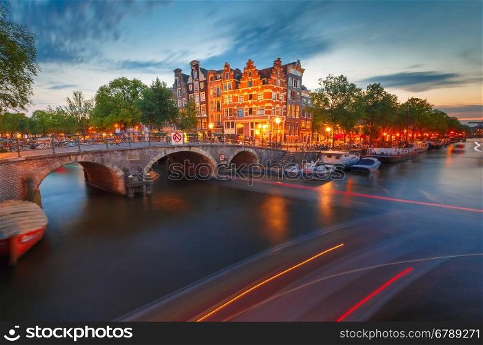 Night city view of Amsterdam canal and bridge. Amsterdam canal, bridge and typical houses, boats and luminous track from the boat during morning twilight blue hour, Holland, Netherlands.