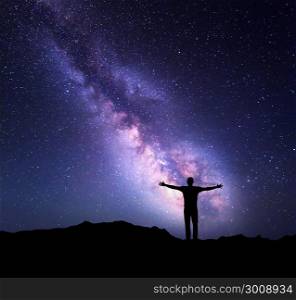 Night colorful landscape with Milky Way and silhouette of a standing sporty man with raised up arms on the mountain. Beautiful Universe. Travel background with purple sky full of stars