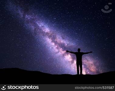 Night colorful landscape with Milky Way and silhouette of a standing sporty man with raised up arms on the mountain. Beautiful Universe. Travel background with purple sky full of stars
