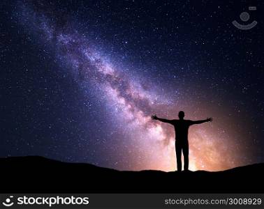 Night colorful landscape with Milky Way, yellow city lights and silhouette of a standing sporty man with raised up arms on the mountain. Beautiful Universe. Travel background with purple sky full of stars