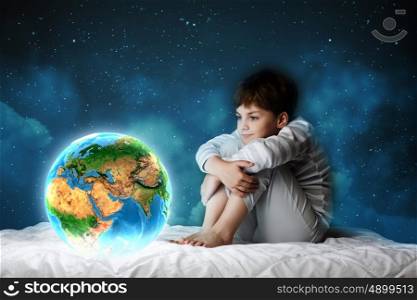 Night dreaming. Cute boy sitting in bed and looking at Earth planet. Elements of this image are furnished by NASA