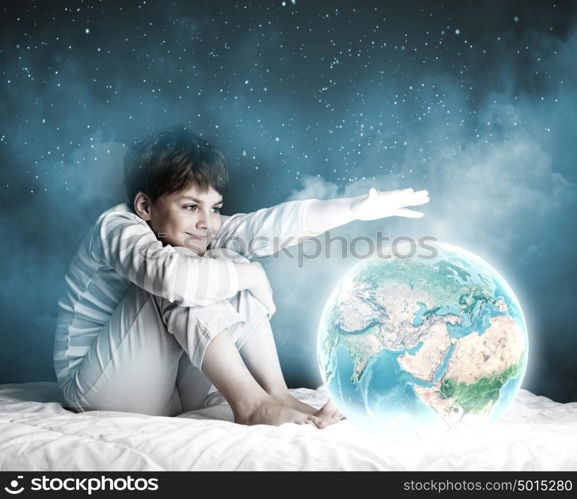 Night dreaming. Cute boy sitting in bed and looking at Earth planet. Elements of this image are furnished by NASA