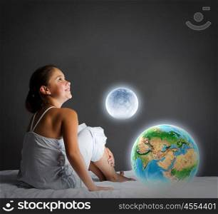 Night dreaming. Cute girl sitting in bed and looking at Earth planet. Elements of this image are furnished by NASA
