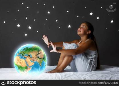 Night dreaming. Cute girl sitting in bed and looking at Earth planet. Elements of this image are furnished by NASA