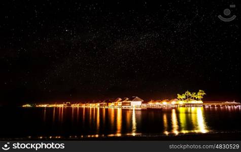 Night on tropical resort, many little beach houses glowing bright yellow lights in dark night, starry sky over sea, romantic summer holidays concept
