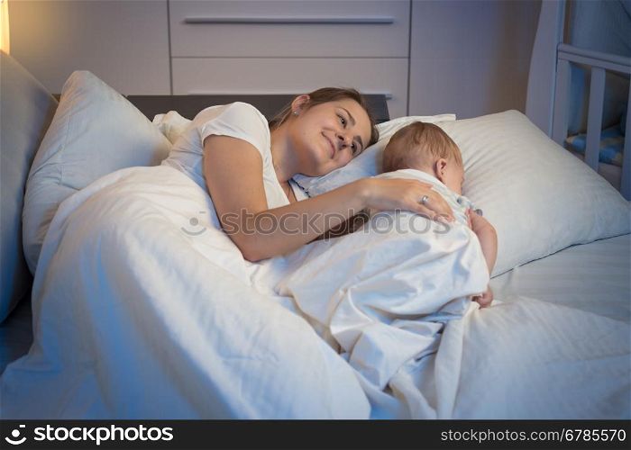 Night shot of happy smiling mother lying on bed with her beautiful baby