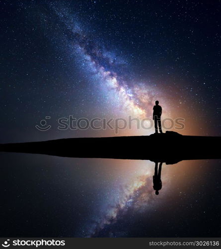 Night starry sky with Milky Way and silhouette of a standing man on the hill near the lake with sky reflection in water. Milky way and man. Galaxy and silhouette of a man. Universe. Travel background