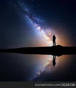 Night starry sky with Milky Way and silhouette of a standing man on the hill near the lake with sky reflection in water. Milky way and man. Galaxy and silhouette of a man. Universe. Travel background
