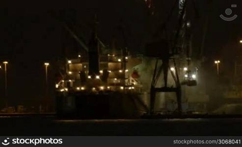Night view of a harbour in a port with an illuminated cargo ship docked at the wharf being offloaded using cranes