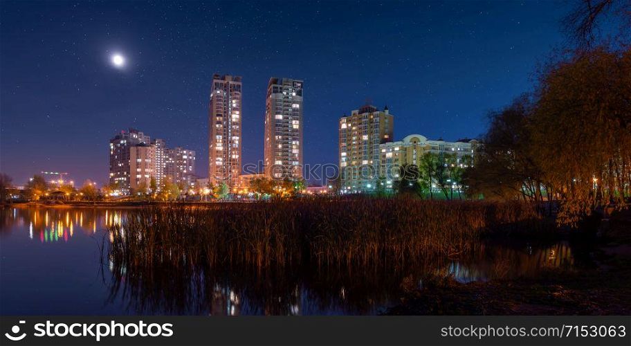 Night view of modern buildings in the obolon district of Kiev, Ukraine, viewed from the southern part of Natalka Park. The moon shines in the starry blue sky. The lights reflect on Dnieper river.