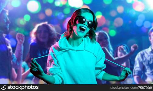 nightlife, entertainment and people concept - happy young woman in black sunglasses wearing hoodie at nightclub over lights background. woman in hoodie and sunglasses at nightclub